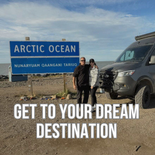 A man and a woman stand in front of a blue 'Arctic Ocean' sign next to a winnebago revel sprinter on a dirt road