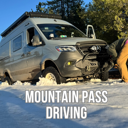 A gray camper van parked in snow with a person bent over near the front bumper, trees and blue sky in the background, and large text reading 'MOUNTAIN PASS DRIVING' across the bottom.