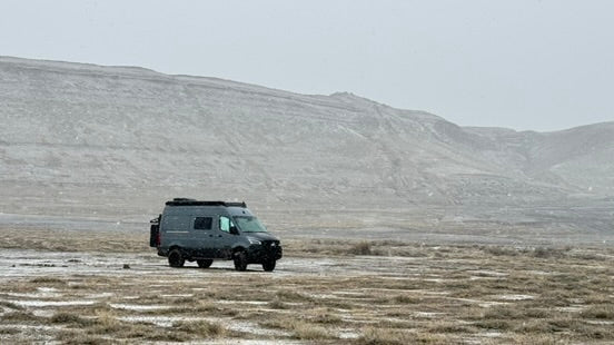 A dark gray camper van with a roof rack is parked on a dry, grassy plain with rocky hills in the background under an overcast sky.